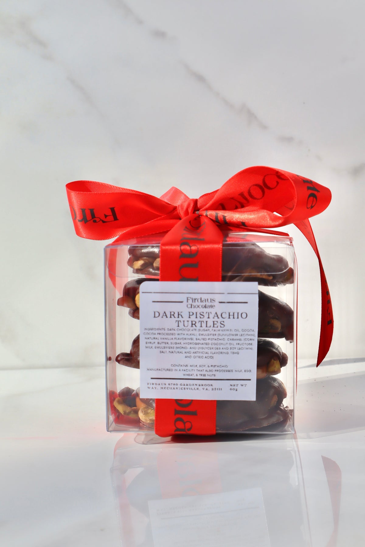 A clear square gift box filled with dark pistachio turtles chocolate, tied with a bright red ribbon and a white product label, set against a white marble background.