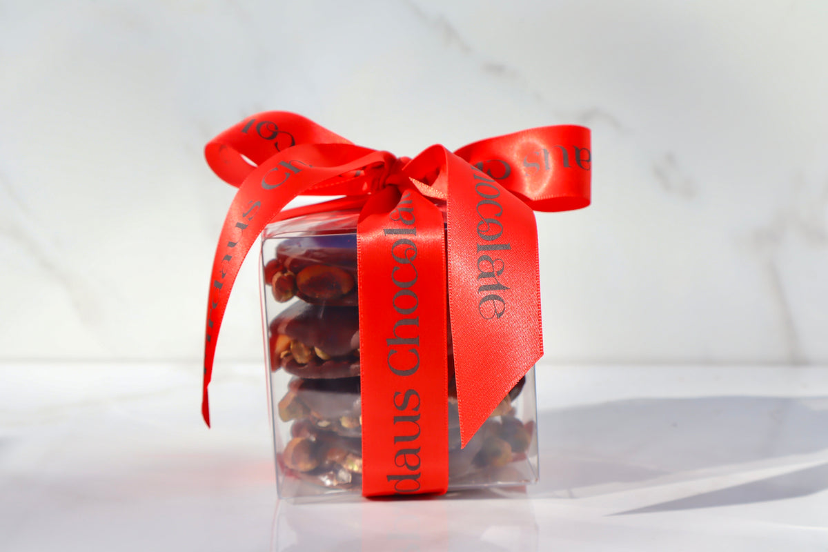 A clear square plastic gift box filled with chocolate pieces, tied with a bright red satin ribbon printed with the words 'Firdaus Chocolate'.